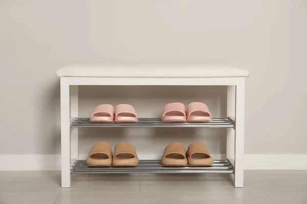 Stylish storage bench with different pairs of rubber slippers near white wall in hall