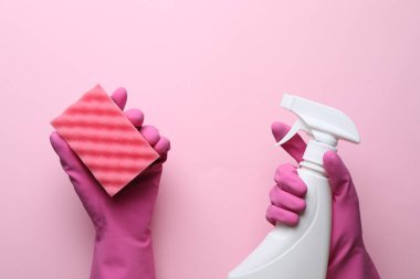 Lastik eldivenli kadın pembe arka planda sünger ve deterjan tutuyor.