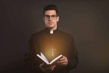 Priest in cassock holding Bible with holy light on black background