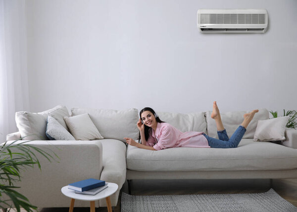 Young woman resting under air conditioner on white wall at home