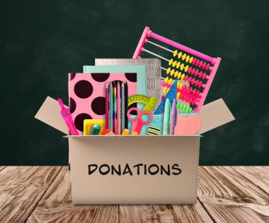 Tahtanın yanındaki ahşap masada farklı okul kırtasiyeleri olan bağış kutusu.