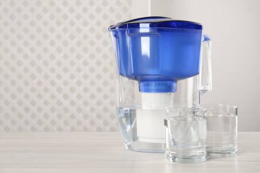 Filter jug and glasses with purified water on white table indoors. Space for text