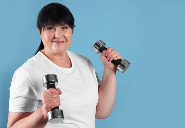 Happy overweight mature woman doing exercise with dumbbells on light blue background