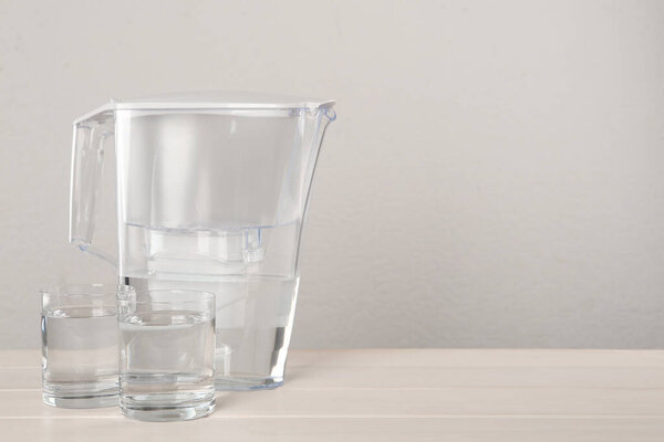 Filter jug and glasses with purified water on white table against light background. Space for text