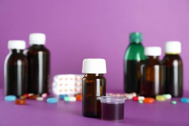 Bir şişe öksürük şurubu ve mor arka planda ölçü kabı.