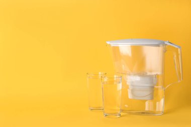 Filter jug and glasses with purified water on yellow background. Space for text