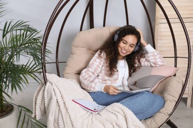 Kulaklık ve tabletle evde yumurta sandalyesinde ders çalışan Afro-Amerikalı bir kadın. Uzaklık öğrenimi