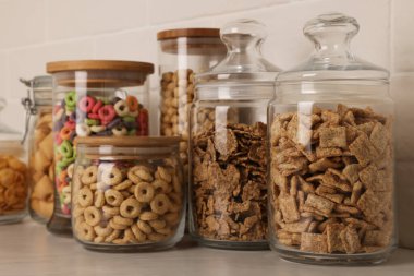 Tuğla duvarın yanındaki beyaz tezgahta farklı kahvaltı gevrekleri olan cam kaplar.