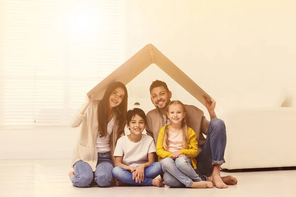 Happy family sitting under cardboard roof at home. Insurance concept