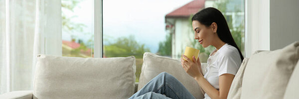 Young woman with cup of coffee relaxing on sofa at home. Banner design