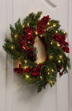 Beyaz kapısında kırmızı böğürtlen ve peri ışıkları asılı güzel Noel çelengi