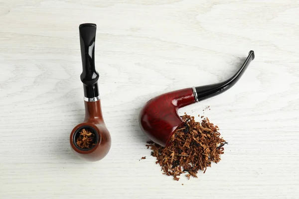 Smoking pipes with tobacco on white wooden table, flat lay