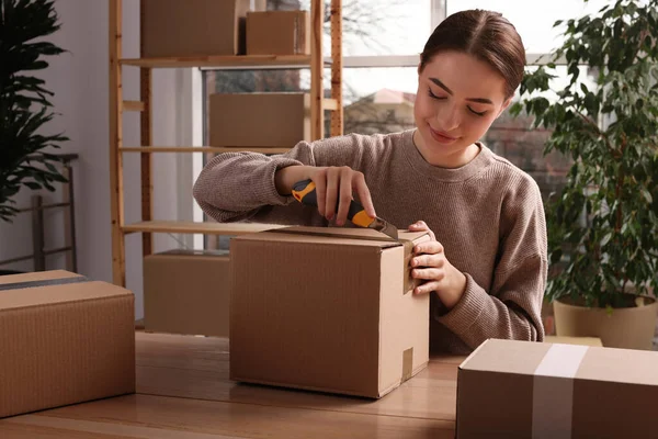 Young Woman Using Utility Knife Open Parcel Wooden Table Indoors — стоковое фото