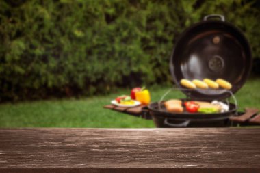 Boş ahşap masa ve modern barbekü ızgarasında leziz yiyeceklerin bulanık görüntüsü.