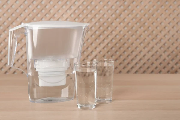 Filter jug and glasses with purified water on wooden table indoors. Space for text