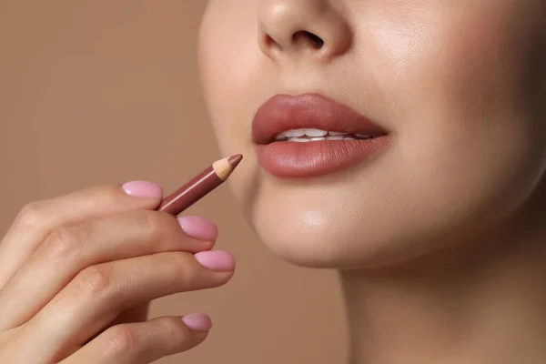 Young woman applying beautiful nude lip pencil on beige background, closeup