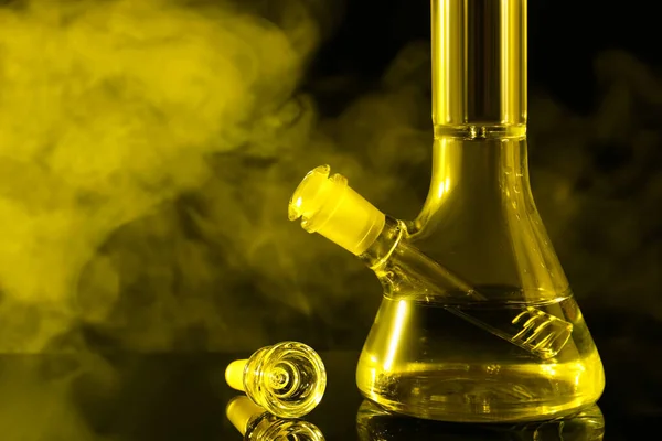 Closeup view of glass bong with smoke on black background, toned in yellow. Smoking device