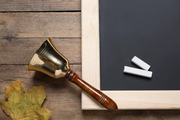 Golden school bell, autumn leaf and blackboard with pieces of chalk on wooden table, flat lay