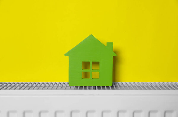 Wooden house model on heating radiator near yellow wall. Energy efficiency concept