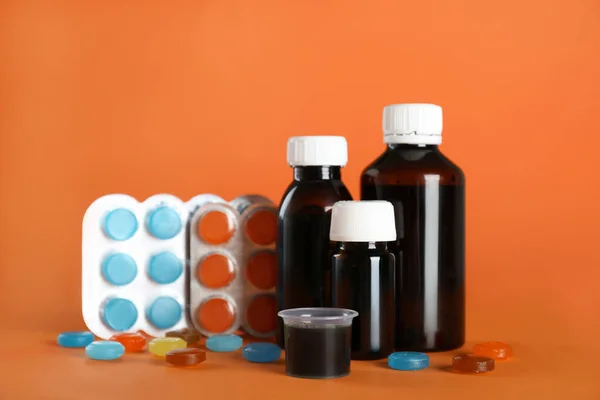 Bottles of syrup, measuring cup and cough drops on orange background