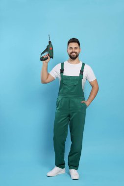 Açık mavi arka planda matkabı olan üniformalı genç bir işçi.