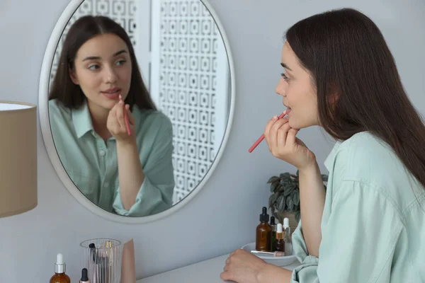 美丽的年轻女子在室内镜子前的唇上涂上化妆品铅笔 — 图库照片