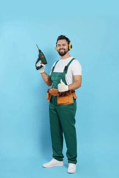 Young worker in uniform with power drill on light blue background