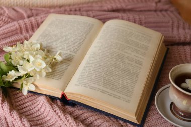 Açık bir kitap, bir fincan aromatik çay ve pembe kumaşta güzel yasemin çiçekleri.