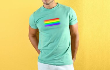 Sarı arka planda LGBT gurur bayrağı resmi olan turkuaz tişört giyen genç adam