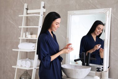 Güzel genç bir kadın banyoda aynanın yanında nemlendirici krem sürüyor.