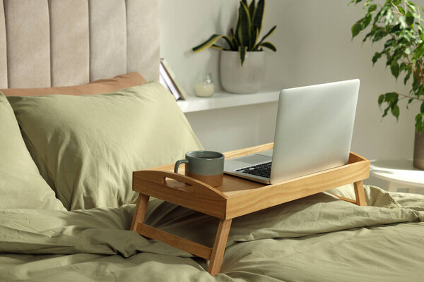 Деревянный поднос с современным ноутбуком и чашкой ароматного чая на кровати в помещении