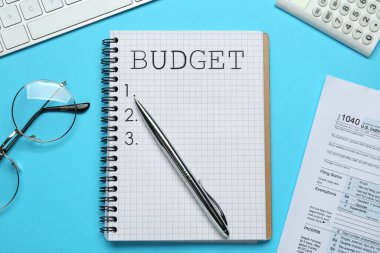 Bütçe, dokümanlar, bilgisayar klavyesi, hesap makinesi ve açık mavi zemin üzerindeki gözlüklü defter, düz konum