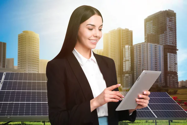 Young businesswoman with tablet near solar panels and beautiful view of cityscape. Alternative energy source