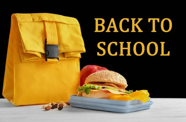 Lunch box with appetizing food and bag on white wooden table near blackboard