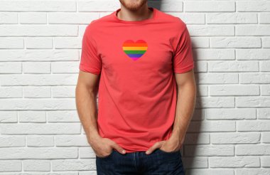 Young man wearing t-shirt with image of heart shaped LGBT pride flag near white brick wall