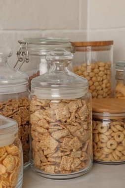 Tuğla duvarın yanındaki beyaz tezgahta farklı kahvaltı gevrekleri olan cam kaplar.