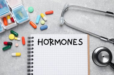 Notebook with word HORMONES, stethoscope and pills on grey table, flat lay