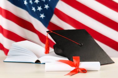 Amerikan bayrağına karşı siyah mezuniyet şapkası, diploma ve beyaz tahta masa üzerine kitap.