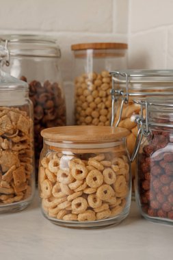 Tuğla duvarın yanındaki beyaz tezgahta farklı kahvaltı gevrekleri olan cam kaplar.