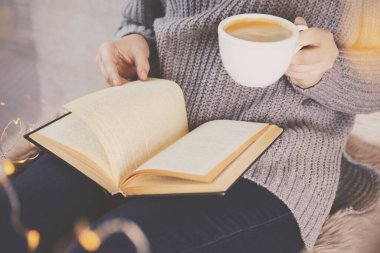 Elinde kahve fincanı olan bir kadın evde kitap okuyor.