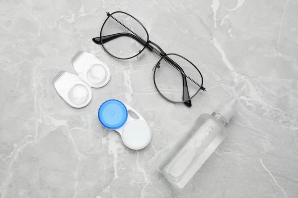 Packages with contact lenses, case, glasses and drops on light grey marble table, flat lay