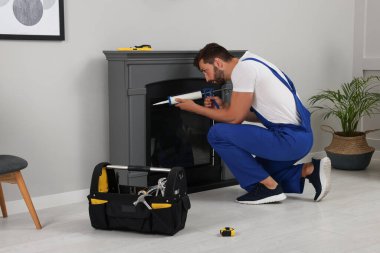 Professional technician sealing electric fireplace with caulk near white wall in room