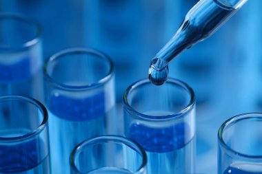 Açık mavi arka planda test tüpüne reaktör damlatılıyor, yakın plan. Laboratuvar analizi