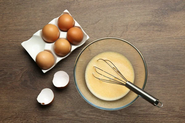 Beaten eggs and whisk on wooden table, flat lay