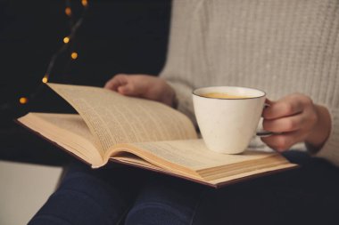 Elinde kahve fincanı olan bir kadın evde kitap okuyor.