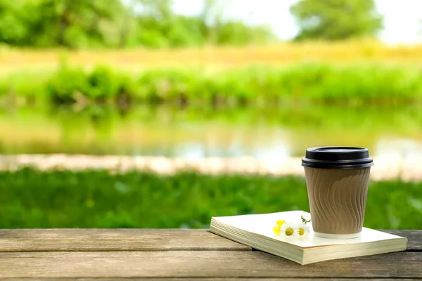 Paper coffee cup, flowers and book on wooden table outdoors, space for text