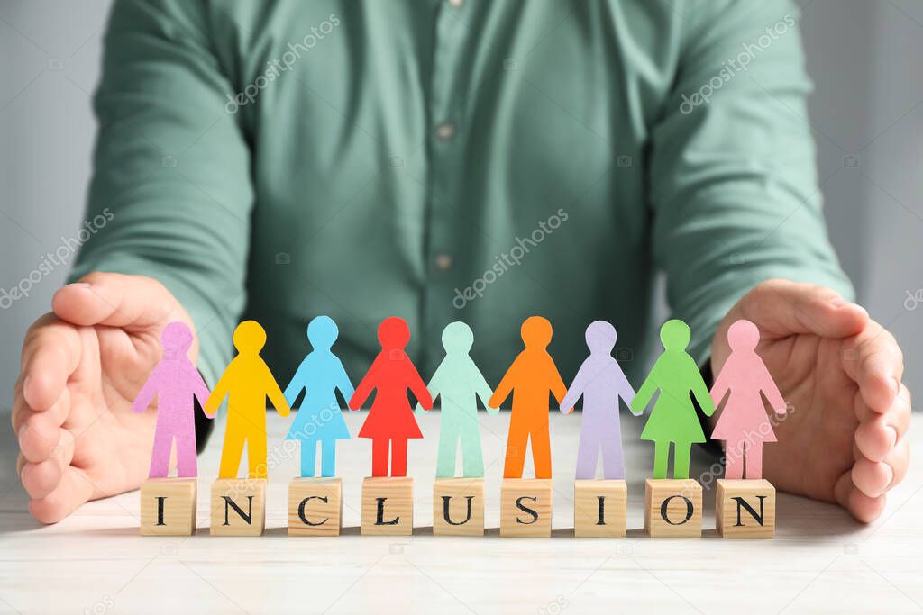 Man protecting paper human figures and wooden cubes with word Inclusion at table indoors, closeup