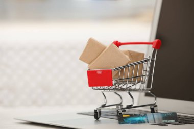 Online payment concept. Small shopping cart with bank card, boxes and laptop on table, space for text