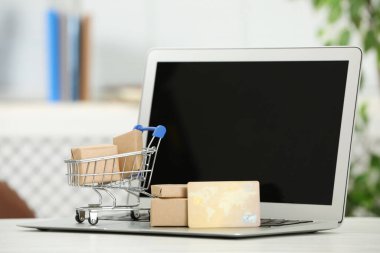 Online payment concept. Small shopping cart with bank card, boxes and laptop on white table