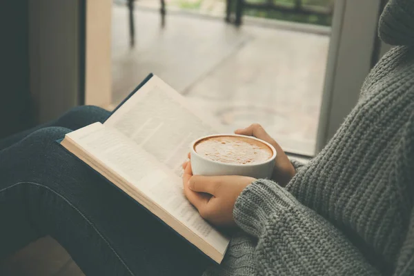 Woman with cup of coffee reading book near window indoors, closeup